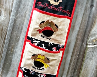 May include: A beige fabric wall organizer with three pockets. Each pocket features a Mickey Mouse head with a pirate hat and a name:  Rooney, Melanie, and Emily. The top pocket has a black fabric panel with the text "Yo Ho Ho The McGee Family" and a small embroidered ship.