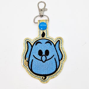 May include: A blue genie character with a gold glitter outline, embroidered on a fabric keychain with a silver clip.