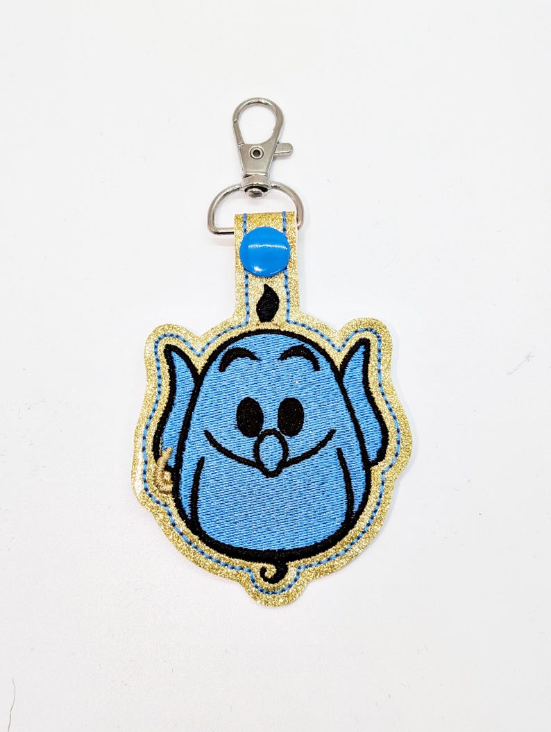 May include: A blue genie character with a gold glitter outline, embroidered on a fabric keychain with a silver clip.