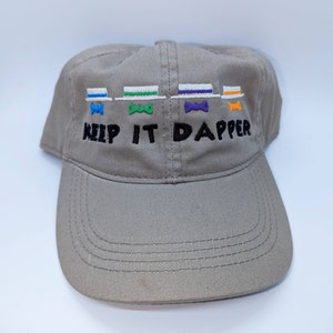 May include: A tan baseball cap with the embroidered phrase "KEEP IT DAPPER." Above the text are four embroidered bow ties and hats in blue, green, purple, and orange. The cap has a curved brim and a structured crown.