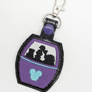 May include: Purple and black glitter keychain with a silhouette of a ride with three people inside. The keychain has a blue Mickey Mouse head on the front.