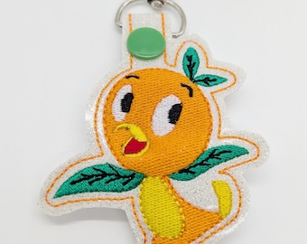 May include: Orange bird character keychain with green leaves, white glitter background, and a silver clip.