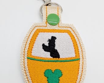 May include: A yellow and green embroidered keychain shaped like a ride vehicle with a silhouette of a cartoon character inside. The keychain has a green Mickey Mouse head on the bottom and a silver key ring.