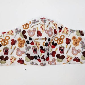 May include: White face mask with an all-over pattern of cartoon food and character designs. The design includes pretzels, pizza slices, ice cream bars, popcorn, and Mickey Mouse-shaped treats. The mask has white adjustable ear loops.