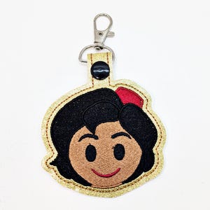 May include: A gold glitter keychain with a black and brown embroidered image of Snow White's face. The keychain has a silver metal clasp.