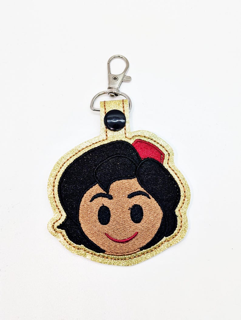 May include: A gold glitter keychain with a black and brown embroidered image of Snow White's face. The keychain has a silver metal clasp.