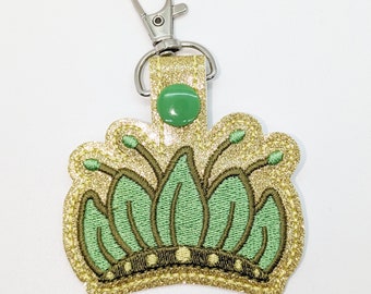 Op de afbeelding: Een sleutelhanger van gouden glitter met een groen kroonontwerp. De kroon heeft een groene edelsteen in het midden en een zilveren sleutelring.