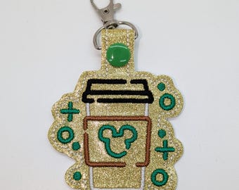 May include: Gold glitter coffee cup keychain with a green Mickey Mouse head inside the cup and green plus and circle designs around the cup. The keychain has a silver metal clip.