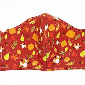 May include: A red fabric face mask with a Thanksgiving theme. The mask features a repeating pattern of turkeys, pumpkins, leaves, and pears.