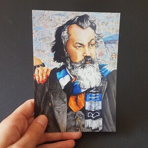 Puede incluir: Una postal con un retrato detallado de un hombre con barba y bigote largos y blancos, que lleva un abrigo negro, una corbata naranja y una bufanda azul y blanca. El fondo muestra una multitud. La postal es sostenida por una mano.