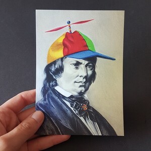 Könnte beinhalten: Eine Postkarte mit einem Porträt eines Mannes, der eine bunte Kappe mit einem Propeller trägt. Die Kappe ist in gelbe, rote, grüne und blaue Abschnitte unterteilt. Der Mann trägt einen dunklen Anzug mit einer Fliege. Der Hintergrund ist hellbeige.