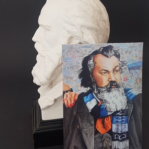 Puede incluir: Un busto de yeso blanco de un hombre con barba larga est&aacute; junto a una postal. La postal presenta un retrato a l&aacute;piz de color de un hombre barbudo, con una bufanda a rayas azules y blancas con la palabra "BRKN".
