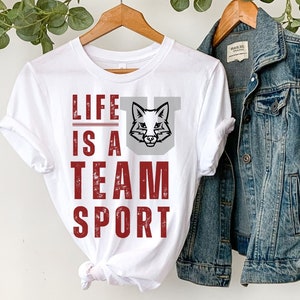 May include: White t-shirt with a red graphic that says "Life is a Team Sport" and a fox head graphic. The shirt is hanging on a rack with a denim jacket and a straw hat.