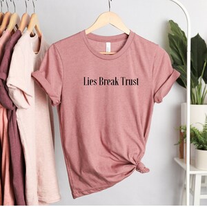May include: A dusty rose colored t-shirt with the text "Lies Break Trust" printed in black on the front.