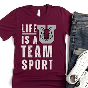 May include: A maroon t-shirt with a distressed white graphic that says "Life is a Team Sport". A gray fox head is in the graphic. The shirt is on a white background with a pair of blue jeans and white sneakers.
