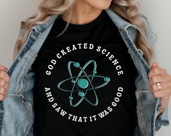 Op de afbeelding: Zwart T-shirt met een witte cirkelvormige afbeelding met een blauw atoom en de tekst "God schiep de wetenschap en zag dat het goed was."