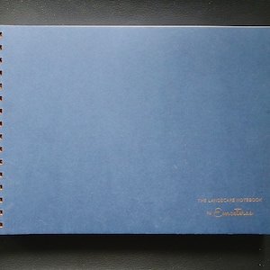May include: A blue notebook with a gold spiral binding. The cover has the text "The Landscape Notebook by Emotinas" printed in gold.