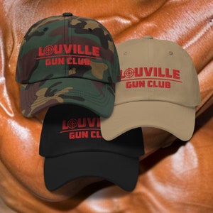 Op de afbeelding: Drie baseballpetten met de tekst "Louisville Gun Club" in rood met een doelwitsymbool. De petten zijn in camouflage, kaki en zwart.