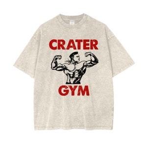 May include: A light beige t-shirt with the words "CRATER GYM" in red, above and below a black illustration of a muscular person flexing their biceps. The shirt has a vintage, worn look.