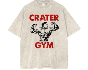 May include: A light beige t-shirt with the words "CRATER GYM" in red, above and below a black illustration of a muscular person flexing their biceps. The shirt has a vintage, worn look.