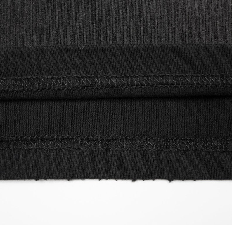 May include: A black fabric garment featuring a double-stitched hem with visible thread detailing. The edge of the fabric is raw and unfinished, showing a textured, slightly frayed appearance along the bottom border of the material.