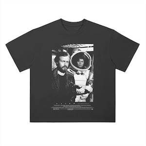 May include: Black and white graphic t-shirt featuring a scene from the movie "Alien". The image shows two characters, one in a spacesuit and the other in a leather jacket. The text "Alien" is printed below the image.