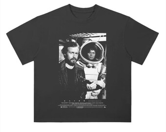 May include: Black and white graphic t-shirt featuring a scene from the movie "Alien". The image shows two characters, one in a spacesuit and the other in a leather jacket. The text "Alien" is printed below the image.