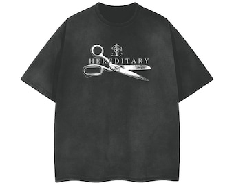 May include: A black short-sleeve t-shirt featuring a white graphic print on the chest. The design displays a pair of open scissors with a small tree icon above them and the word HEREDITARY printed in white capital letters across the center.