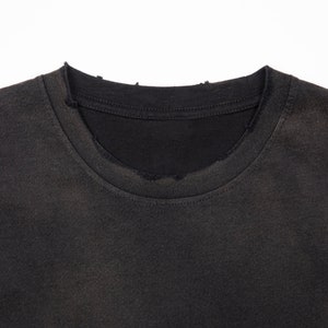 May include: A charcoal black t-shirt featuring a crew neckline with visible distressing and fraying along the collar edge. The fabric has a faded, worn-in appearance with a consistent dark gray tone throughout the visible portion of the garment.