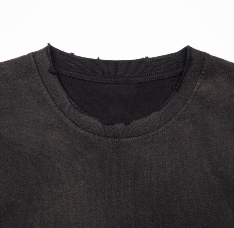 May include: A charcoal black t-shirt featuring a crew neckline with visible distressing and fraying along the collar edge. The fabric has a faded, worn-in appearance with a consistent dark gray tone throughout the visible portion of the garment.