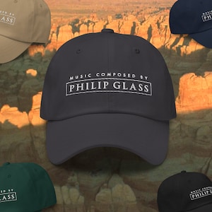 Può includere: Un cappello da baseball nero con testo bianco che recita "Musica composta da Philip Glass".