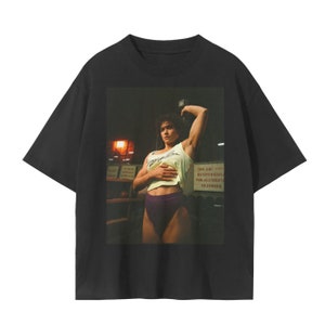 May include: Black t-shirt with a graphic print of a woman lifting weights in a gym. The woman is wearing a white tank top with the text "Angel Love" and purple athletic shorts. The text "We are not responsible for accidents or injuries" is visible on a sign in the background.
