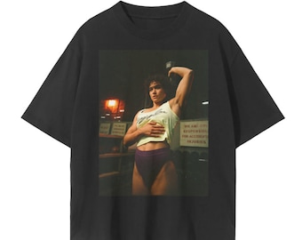 May include: Black t-shirt with a graphic print of a woman lifting weights in a gym. The woman is wearing a white tank top with the text "Angel Love" and purple athletic shorts. The text "We are not responsible for accidents or injuries" is visible on a sign in the background.
