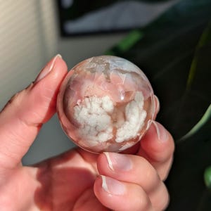 May include: A clear, spherical gemstone with a delicate pattern of pink and white cloud-like formations. The polished surface reflects light, showcasing the natural beauty of the stone. This decorative sphere is a unique addition to any crystal collection.