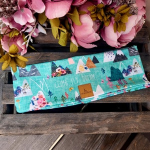Könnte beinhalten: Ein türkisfarbenes Stirnband mit einem Berg- und Blumenmuster. Das Stirnband trägt den Text "Climb Fly Dream" und ein kleines braunes Leder-Etikett mit dem Text "Heimatgfaß".