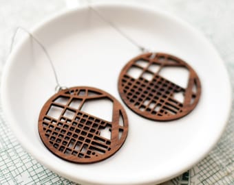 May include: A pair of wooden earrings with a geometric design, resting on a white ceramic dish. The earrings are circular with a cut-out pattern of lines and squares. The earrings are brown and have silver-colored hooks.