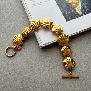 Op de afbeelding: Een goudkleurige armband met schelpvormige schakels. De armband heeft een tuimelsluiting en een cirkelvormige ring. De armband wordt getoond op een boek met tekst en een schilderij.