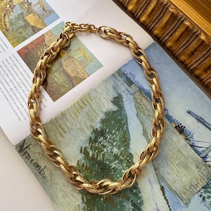 May include: A gold-tone chain necklace featuring thick, interlocking oval links with a textured, cross-hatched surface pattern. The necklace includes a sturdy metal clasp and a short extender chain at the back for adjustable length.