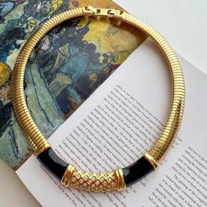 May include: Gold-tone collar necklace with a ribbed design. The necklace features black enamel accents and a central section adorned with clear crystals. A vintage-inspired statement piece.