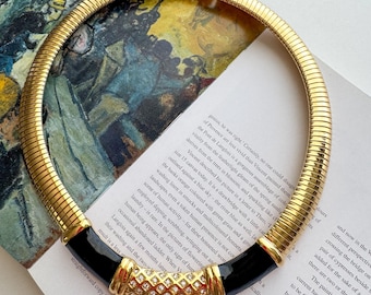 May include: Gold-tone collar necklace with a ribbed design. The necklace features black enamel accents and a central section adorned with clear crystals. A vintage-inspired statement piece.