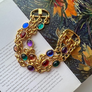 May include: A gold-tone multi-strand chain bracelet featuring oval-shaped cabochon stones in red, blue, green, and purple. The bracelet includes two gold-tone buckle-style clasps at each end and is engraved with the text Kate Spade New York.