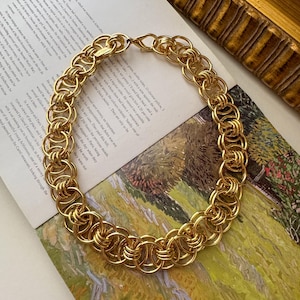 May include: A gold-toned chain necklace featuring a series of interlocking circular links. The necklace is constructed with a repeating pattern of multiple rings connected together to form a continuous, thick, and textured circular chain design.