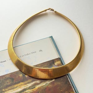 May include: A gold-toned collar necklace with a ribbed design. The necklace is circular and lies on a book with text that reads "of an Endless Whole". The background features a painting with brown and green tones.