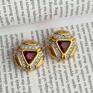 Può includere: Un paio di orecchini dorati con un design geometrico. Ogni orecchino presenta una pietra triangolare rosso scuro al centro, circondata da cristalli chiari e scintillanti e un bordo dorato. Gli orecchini sono posti su uno sfondo di testo stampato.