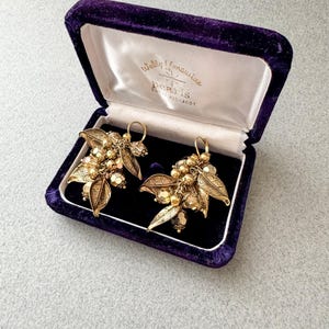 May include: A pair of vintage gold-toned earrings with leaf and bead details, presented in a purple velvet jewelry box. The box is open, revealing the earrings against a black interior. The box has the text "Wally Yomanuise Pearls".