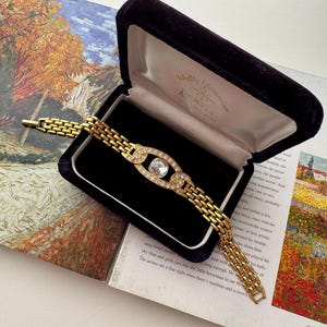 May include: A gold-tone link bracelet featuring a central oval-shaped centerpiece set with a single clear stone surrounded by smaller clear stones. The bracelet is displayed in a dark purple velvet-lined jewelry box with a hinged lid.