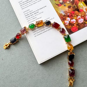 May include: A gold bracelet featuring a series of multi-coloured, rectangular gemstones. The gemstones are in shades of red, green, yellow, and blue. The bracelet is displayed on a book page with text and a painting.