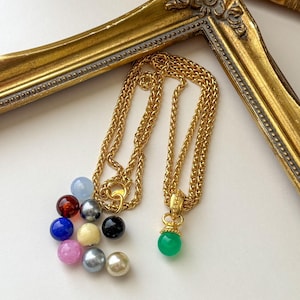 May include: A gold-toned chain necklace featuring a single green spherical pendant. The necklace is accompanied by a collection of loose, multi-colored spherical beads in shades of blue, red, black, white, pink, and grey.