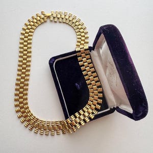 Könnte beinhalten: Eine goldfarbene Metallgliederkette mit einem sich wiederholenden, strukturierten Muster aus horizontalen Balken. Die Kette liegt neben einer geöffneten, rechteckigen Schmuckschatulle mit dunkelviolettem Samtinnenfutter und weißem Satin.