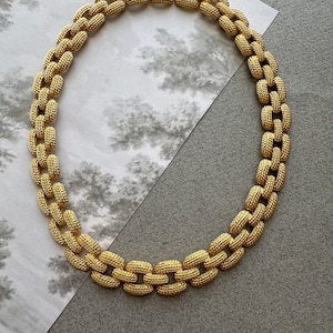 May include: A gold-toned necklace featuring a series of interlocking oval links. Each individual link has a textured, cross-hatched surface finish. The necklace is secured with a small, integrated rectangular clasp at the top center of the chain.